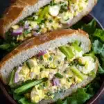 Egg salad sandwich baguette - the image shows two sandwiches in a brown bowl. the sandwiches are made with two slices of toasted bread and are filled with scrambled eggs, green onions, and red onions. the eggs are yellow and runny, and the green onions are scattered throughout the sandwich. the bowl is garnished with a sprig of parsley. the background is dark, making the sandwiches stand out.