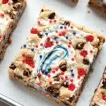 Fourth of july cookie bar - the image shows a tray of freshly baked chocolate chip cookie bars. the bars are rectangular in shape and have a golden brown color. they are topped with a swirl of white frosting in the shape of a red, white, and blue swirl. the frosting is drizzled over the top of the bars, creating a swirl pattern. there are also small red and blue sprinkles scattered throughout the frosting. the tray is lined with parchment paper and there are a few more bars visible in the background.
