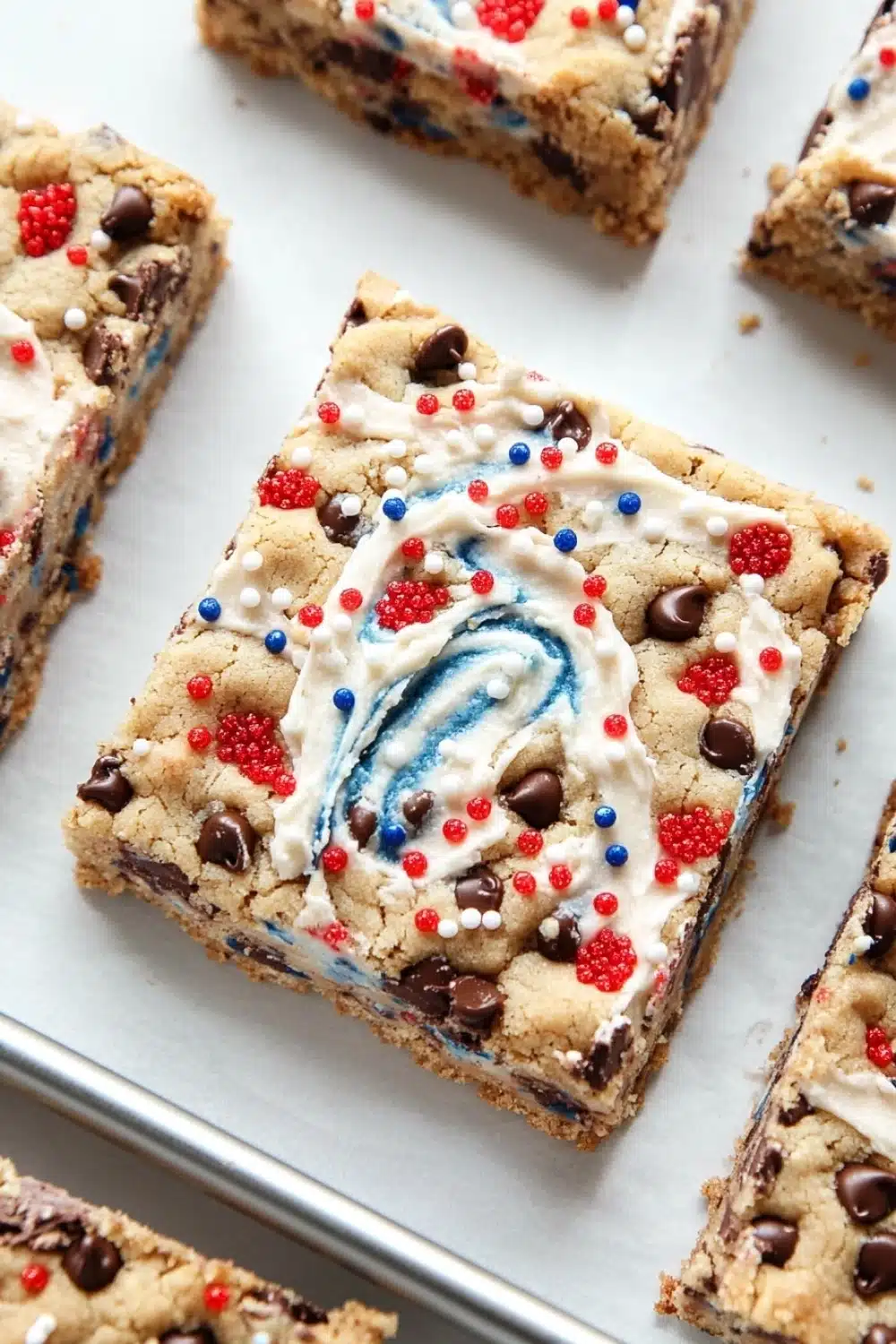 Fourth of july cookie bar - the image shows a tray of freshly baked chocolate chip cookie bars. the bars are rectangular in shape and have a golden brown color. they are topped with a swirl of white frosting in the shape of a red, white, and blue swirl. the frosting is drizzled over the top of the bars, creating a swirl pattern. there are also small red and blue sprinkles scattered throughout the frosting. the tray is lined with parchment paper and there are a few more bars visible in the background.