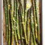 Garlic parmesan roasted asparagus - the image shows a baking tray filled with cooked asparagus spears. the spears are arranged in a single layer on the tray, with some of them overlapping each other. the tray is lined with parchment paper and there are small pieces of lemon wedges scattered on top. the background is a light beige color, and the tray appears to be sitting on a white countertop.