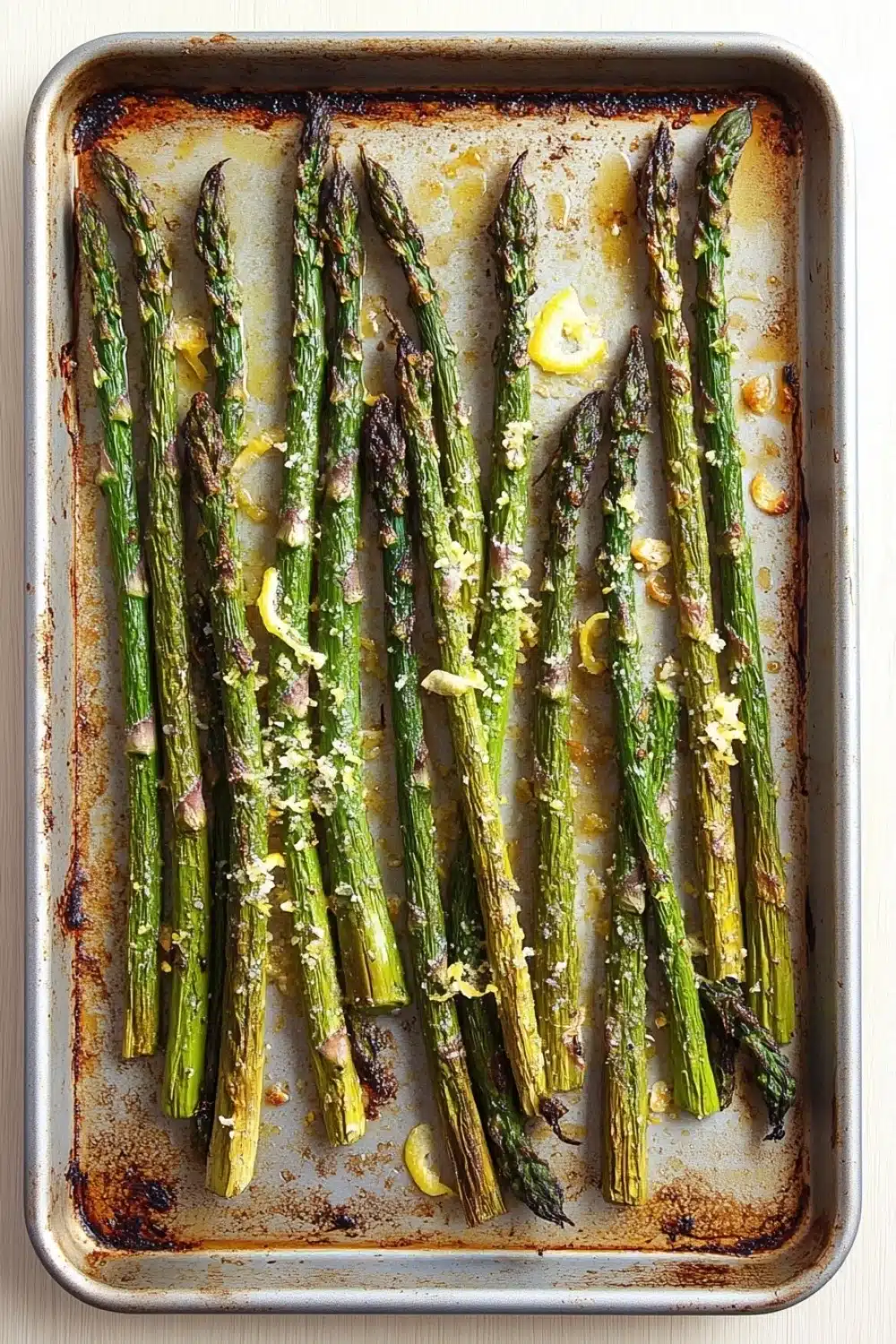 Garlic parmesan roasted asparagus - the image shows a baking tray filled with cooked asparagus spears. the spears are arranged in a single layer on the tray, with some of them overlapping each other. the tray is lined with parchment paper and there are small pieces of lemon wedges scattered on top. the background is a light beige color, and the tray appears to be sitting on a white countertop.
