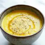Ginger turmeric cauliflower soup - the image is a close-up of a bowl of soup. the bowl is made of ceramic and has a speckled texture. the soup appears to be a creamy yellow color and is topped with a drizzle of orange sauce. the sauce is drizzled over the top of the soup, creating a smooth and creamy texture. there are small black speckles scattered throughout the sauce, adding a pop of color to the dish. the background is white, making the bowl and sauce stand out.