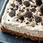 Graham bar cookie and cream - the image is of a slice of oreo cheesecake on a black slate tray. the cheesecake has a creamy white frosting on top, with small oreo cookies scattered throughout. the cookies are arranged in a circular pattern, with some overlapping each other. the tray is lined with parchment paper, and the background is blurred, making the cheesecake the focal point of the image.