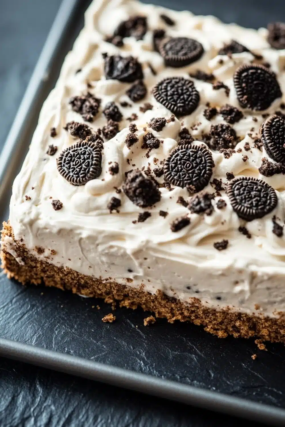 Graham bar cookie and cream - the image is of a slice of oreo cheesecake on a black slate tray. the cheesecake has a creamy white frosting on top, with small oreo cookies scattered throughout. the cookies are arranged in a circular pattern, with some overlapping each other. the tray is lined with parchment paper, and the background is blurred, making the cheesecake the focal point of the image.