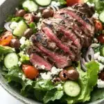 Greek salad with lamb - the image is a close-up of a salad in a gray bowl. the salad is made up of various greens, including lettuce, cherry tomatoes, cucumbers, and red onions. on top of the greens, there is a large piece of grilled steak, which appears to be seasoned with herbs and spices. the steak is cooked to a medium-rare and has a pinkish-red color. there are also small pieces of feta cheese scattered throughout the salad. the bowl is sitting on a white marble countertop.