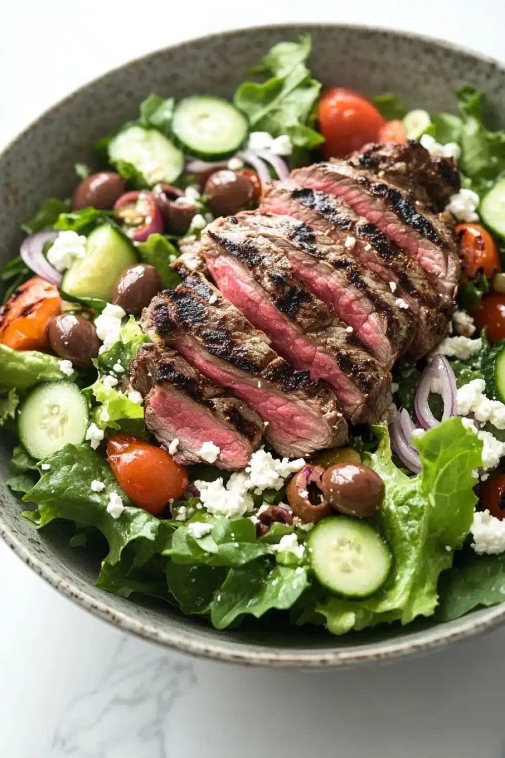Greek salad with lamb - the image is a close-up of a salad in a gray bowl. the salad is made up of various greens, including lettuce, cherry tomatoes, cucumbers, and red onions. on top of the greens, there is a large piece of grilled steak, which appears to be seasoned with herbs and spices. the steak is cooked to a medium-rare and has a pinkish-red color. there are also small pieces of feta cheese scattered throughout the salad. the bowl is sitting on a white marble countertop.