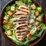 Grilled chicken caesar salad - the image is a close-up of a bowl of caesar salad. the salad is made up of lettuce leaves, croutons, and grilled chicken. the chicken is cooked to a golden brown color and appears to be seasoned with herbs and spices. the lettuce leaves are a vibrant green color and are arranged in a circular pattern around the chicken. there is a sprinkle of white cheese on top of the salad, adding a pop of color to the dish. the bowl is made of dark brown ceramic and is placed on a wooden table.