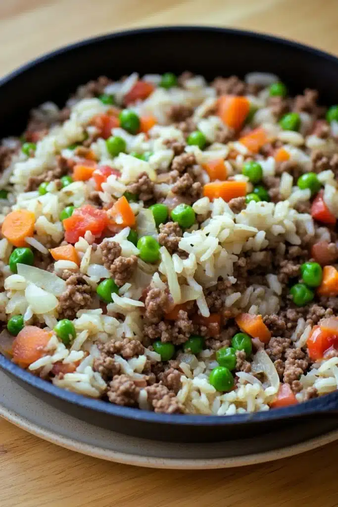 Ground beef and rice - the image shows a close-up of a dish in a black frying pan. the dish appears to be a type of fried rice with ground beef, peas, carrots, and onions mixed in. the rice is white and fluffy, and the ground beef is a dark brown color. the peas are bright green and are scattered throughout the dish. the carrots are bright orange and appear to be diced into small pieces. the onions are light brown and are also diced. the pan is sitting on a wooden table.