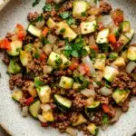 Ground beef and zucchini - the image is a close-up of a plate of food. the plate is white with a speckled pattern and is placed on a wooden table. the food appears to be a salad with a variety of vegetables, including zucchini, bell peppers, onions, and ground beef. the vegetables are mixed in with the ground beef, creating a colorful and appetizing dish. there are also some chopped parsley scattered around the plate, adding a pop of green color to the dish.