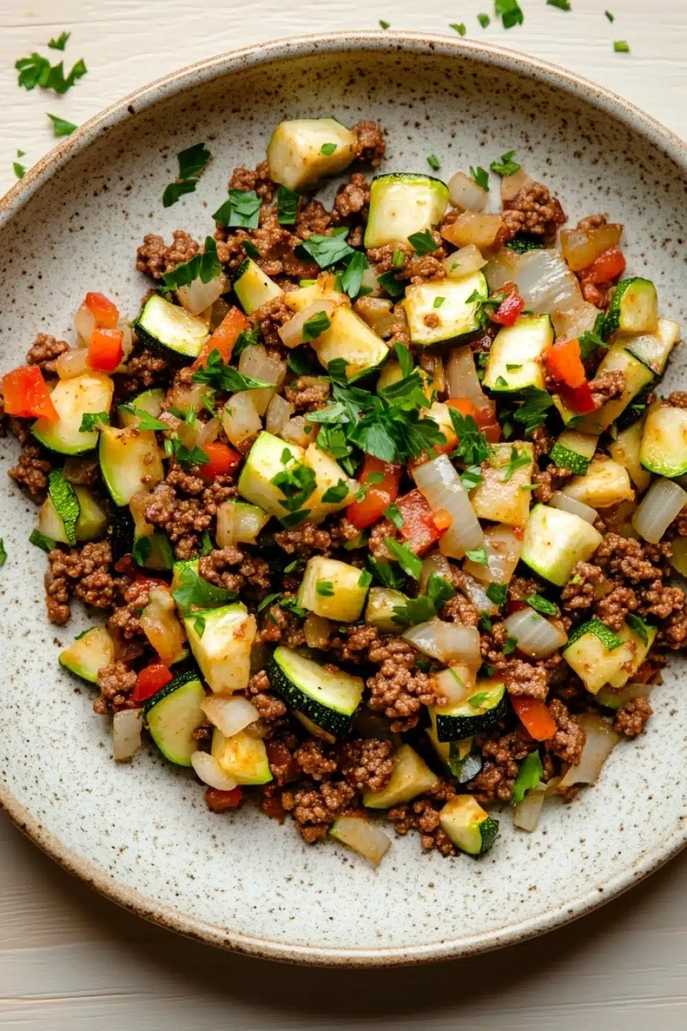 Ground beef and zucchini - the image is a close-up of a plate of food. the plate is white with a speckled pattern and is placed on a wooden table. the food appears to be a salad with a variety of vegetables, including zucchini, bell peppers, onions, and ground beef. the vegetables are mixed in with the ground beef, creating a colorful and appetizing dish. there are also some chopped parsley scattered around the plate, adding a pop of green color to the dish.