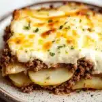 Ground beef casserole with potatoes and cheese - the image shows a plate of food with a layered dish on it. the dish appears to be a shepherd's pie, with a layer of ground beef and potatoes on top. the top of the dish is covered in melted cheese, which is golden brown and bubbly. the cheese is melted and oozing out of the sides. the potatoes are cut into small cubes and are arranged in a pile on the plate. the plate is white with a speckled pattern and there are a few sprigs of parsley scattered around the plate, adding a pop of green color to the dish.