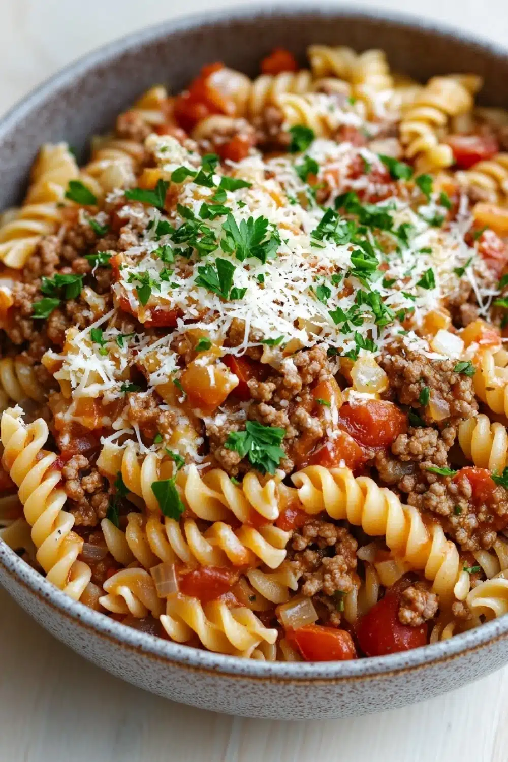 Ground beef with pasta - the image is a close-up of a bowl of pasta dish. the pasta appears to be fusilli, which is a type of spiral-shaped pasta. the dish is made with ground beef, tomato sauce, and chunks of red bell peppers. the beef is cooked in a tomato-based sauce and is topped with grated parmesan cheese. the bowl is made of ceramic and is sitting on a white tablecloth. there are also a few sprigs of fresh parsley scattered on top of the dish, adding a pop of green color.