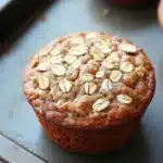 High protein banana oat muffin - the image shows a close-up of a freshly baked muffin on a baking tray. the muffin is golden brown in color and has a crumbly texture. it is topped with sliced oats, giving it a crunchy appearance. the tray is lined with parchment paper and there are other muffins visible in the background. the background is blurred, but it appears to be a kitchen countertop.