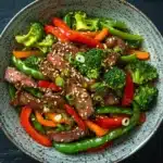 High protein beef stir fry - the image is a close-up of a bowl of stir-fry with beef and vegetables. the bowl is made of ceramic and has a dark blue speckled pattern. the beef is sliced into thin strips and is topped with sesame seeds. the vegetables include broccoli, red bell peppers, and green bell peppers. there are also some sliced carrots and broccoli florets scattered throughout the dish. the dish is garnished with fresh cilantro leaves. the background is a dark grey textured surface.