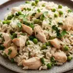 High protein chicken and rice - the image shows a plate of rice with chicken and peas. the rice is white and fluffy, and the chicken is cooked to a golden brown color. the peas are bright green and are scattered throughout the dish. the dish is garnished with a sprig of fresh parsley. the plate is brown and has a textured surface. the background is a white wooden table.
