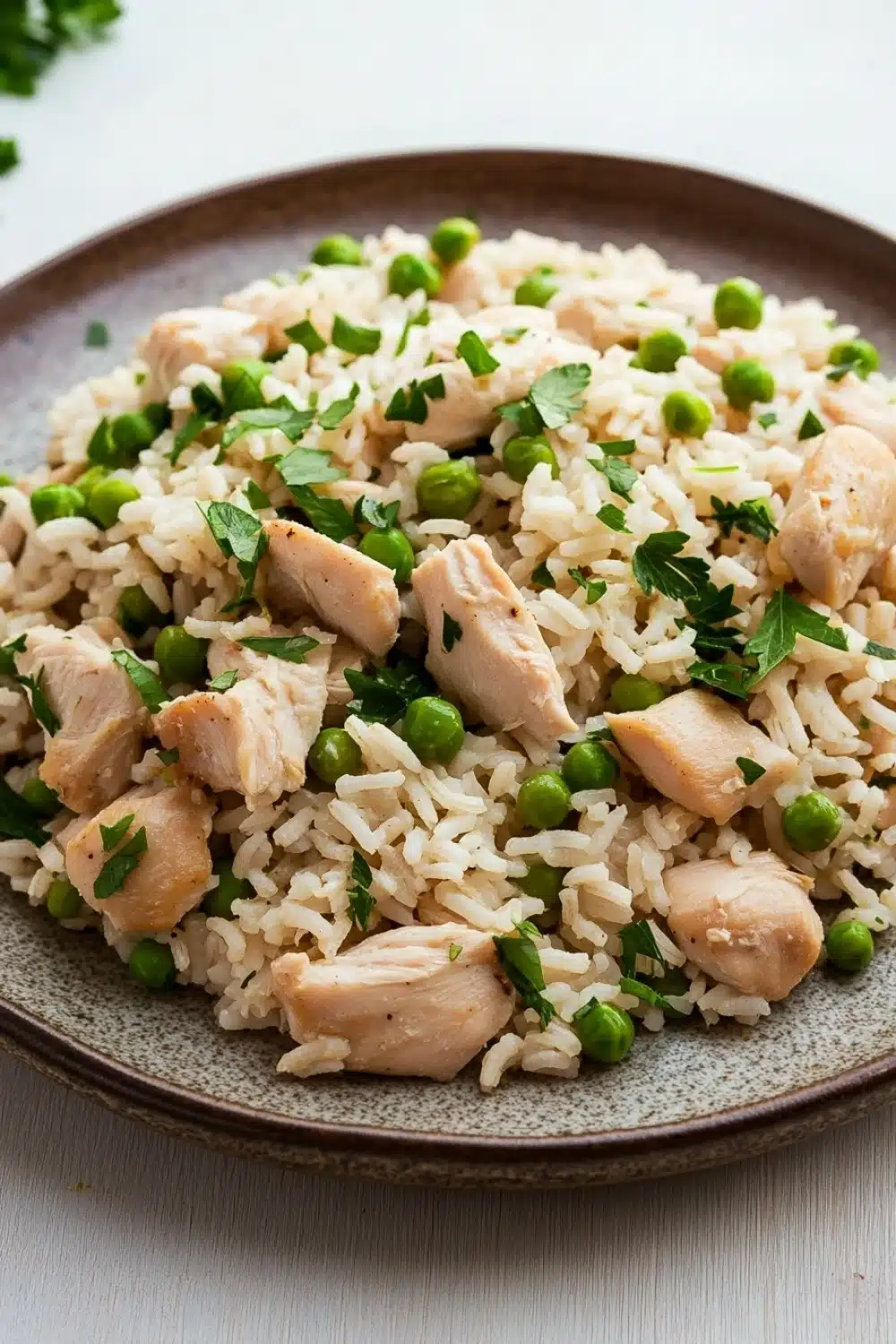 High protein chicken and rice - the image shows a plate of rice with chicken and peas. the rice is white and fluffy, and the chicken is cooked to a golden brown color. the peas are bright green and are scattered throughout the dish. the dish is garnished with a sprig of fresh parsley. the plate is brown and has a textured surface. the background is a white wooden table.