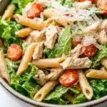 High protein chicken caesar pasta salad - the image is a close-up of a bowl of pasta salad. the bowl is made of ceramic and has a light brown color. the pasta is long and thin, and it appears to be penne. the salad is made up of mixed greens, including lettuce, cherry tomatoes, and crumbled parmesan cheese. there are also chunks of chicken mixed in with the pasta. the overall appearance of the salad is fresh and appetizing.