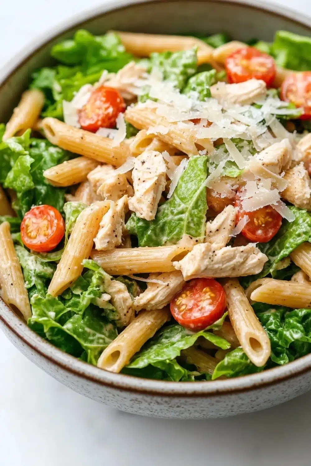 High protein chicken caesar pasta salad - the image is a close-up of a bowl of pasta salad. the bowl is made of ceramic and has a light brown color. the pasta is long and thin, and it appears to be penne. the salad is made up of mixed greens, including lettuce, cherry tomatoes, and crumbled parmesan cheese. there are also chunks of chicken mixed in with the pasta. the overall appearance of the salad is fresh and appetizing.