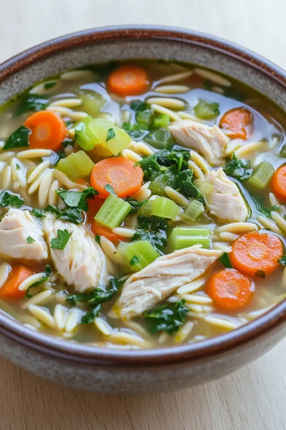 High protein chicken orzo soup - the image is a close-up of a bowl of soup. the soup appears to be a type of noodle soup with chunks of chicken, carrots, celery, and other vegetables mixed in. the broth is a light yellow color and looks thick and creamy. the bowl is made of ceramic and is sitting on a wooden table. the vegetables are bright orange and appear to be fresh and vibrant. there are also some green herbs scattered throughout the soup, adding a pop of color to the dish.