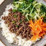 High protein korean beef rice bowl - the image is a close-up of a bowl of food. the bowl is made of a light-colored ceramic material and has a brown rim. on top of the bowl, there is a bed of white rice with chunks of ground beef and green onions scattered on top. on the right side of the rice, there are a few pieces of sliced carrots with sesame seeds sprinkled on top, adding a pop of color to the dish. the dish is garnished with a sprinkle of sesame seed. the background is a wooden table.