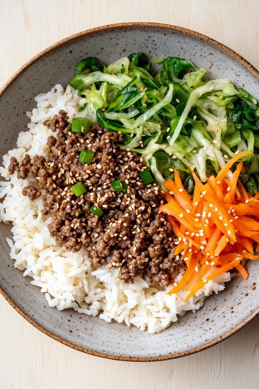 High protein korean beef rice bowl - the image is a close-up of a bowl of food. the bowl is made of a light-colored ceramic material and has a brown rim. on top of the bowl, there is a bed of white rice with chunks of ground beef and green onions scattered on top. on the right side of the rice, there are a few pieces of sliced carrots with sesame seeds sprinkled on top, adding a pop of color to the dish. the dish is garnished with a sprinkle of sesame seed. the background is a wooden table.