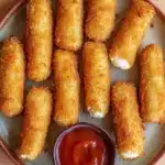 Homemade glutenfree mozzarella sticks - the image shows a plate of deep-fried breaded chicken tenders. the tenders are golden brown and appear to be crispy on the outside. they are arranged in a circular pattern on the plate, with a small bowl of ketchup on the side. the plate is sitting on a wooden table.