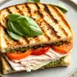 Italian sandwich panini - the image shows a sandwich on a blue and white plate. the sandwich is made with two slices of toasted bread, which are golden brown and appear to be seasoned with herbs and spices. on top of the bread, there is a generous amount of sliced ham, sliced tomatoes, and a dollop of pesto sauce. there is also a sprig of fresh basil on the plate, adding a pop of green color to the dish. the plate is sitting on a white marble countertop.