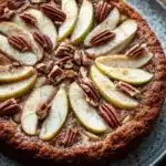 Keto german apple cake - the image is of a round cake with a golden brown crust on a blue plate. the cake is topped with sliced pears and pecans, giving it a crunchy texture. the pears are arranged in a circular pattern on top of the cake, with some overlapping each other. the plate is sitting on a wooden table, and the background is blurred, making the cake the focal point of the image.