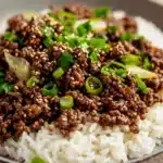 Korean beef skillet - the image shows a plate of food with a serving of rice and ground beef. the rice is white and fluffy, and the ground beef is a dark brown color. the dish is garnished with chopped green onions and sesame seeds. the plate is gray and has a speckled pattern around the edges. the background is blurred, but it appears to be a wooden table.