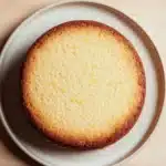 Lemon cake layer - the image is a close-up of a round cake on a white plate. the cake appears to be freshly baked and has a golden brown crust on top. the plate is placed on a light-colored wooden surface. the background is blurred, making the cake the focal point of the image.