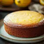 Lemon cake low sugar - the image shows a round cake on a white plate with a blue rim. the cake appears to be a lemon cake with a golden brown crust and a light dusting of powdered sugar on top. in the background, there are three whole lemons and a beige cloth napkin. the table is made of dark wood and the background is blurred, making the cake the focal point of the image.