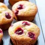 Lemon cherry muffin - the image shows a muffin tin filled with freshly baked cherry muffins. the muffins are golden brown and have a crumbly texture. they are topped with fresh cherries, which are a deep red color and appear to be ripe and juicy. the tin is lined with parchment paper, and the background is blurred, making the muffins the focal point of the image.