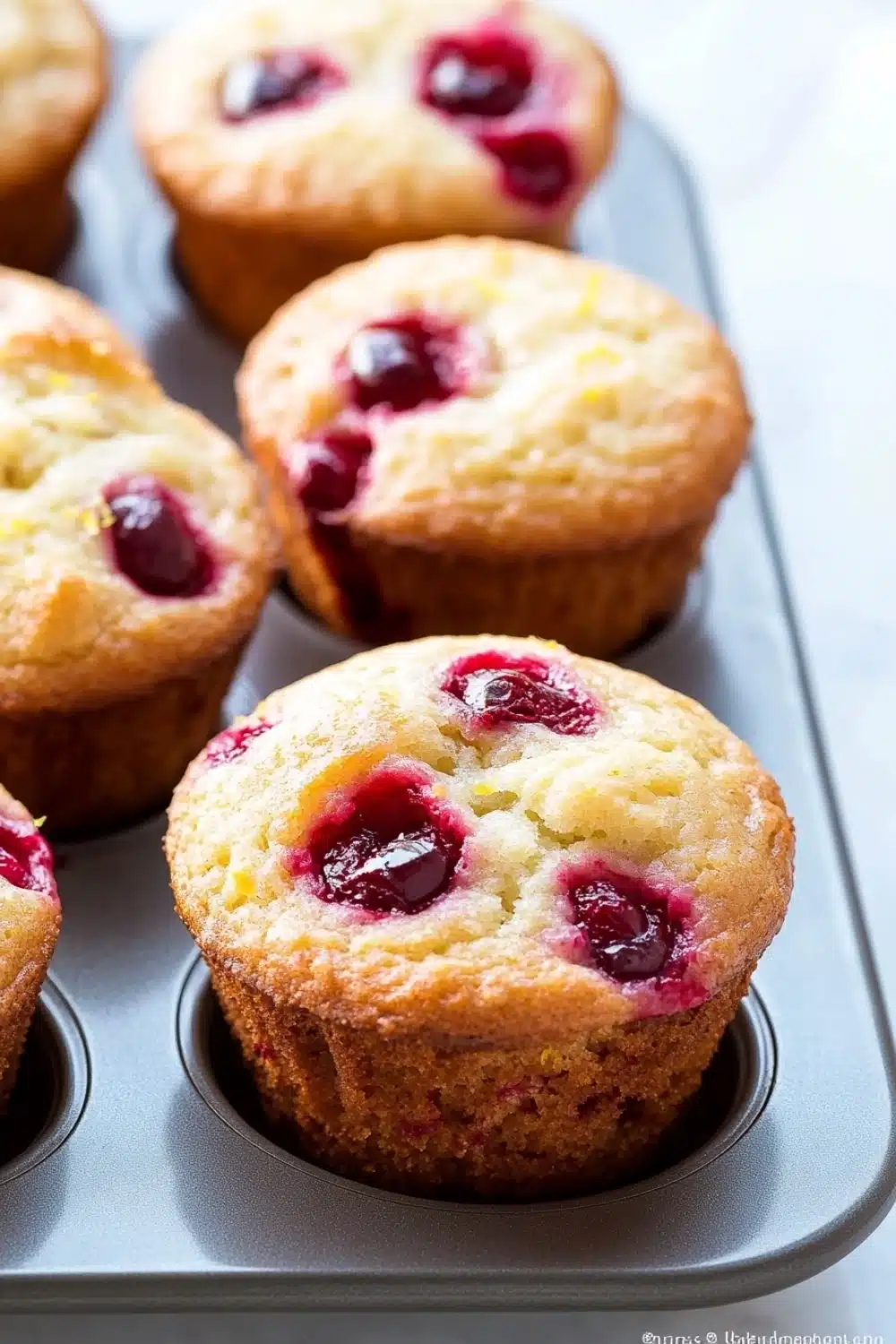 Lemon cherry muffin - the image shows a muffin tin filled with freshly baked cherry muffins. the muffins are golden brown and have a crumbly texture. they are topped with fresh cherries, which are a deep red color and appear to be ripe and juicy. the tin is lined with parchment paper, and the background is blurred, making the muffins the focal point of the image.