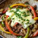 Low carb philly cheese steak skillet with ground beef - the image is a close-up of a dish in a brown and white speckled bowl. the dish appears to be a stir-fry with ground beef, red and yellow bell peppers, onions, and a poached egg on top. the egg is covered in a layer of melted cheese and is garnished with fresh parsley. the bowl is sitting on a wooden table with a white cloth napkin in the background.