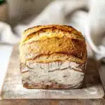 Low carb yeast sandwich bread - the image shows a loaf of bread on a wooden cutting board. the bread appears to be freshly baked and has a golden brown crust on top. the loaf is sitting on a white marble countertop with a white cloth napkin and a potted plant in the background. the overall aesthetic of the image is rustic and homey.
