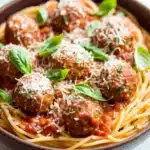 Make ahead pasta and meatballs - the image is a close-up of a bowl of spaghetti and meatballs. the spaghetti is cooked al dente and is covered in a red tomato sauce with chunks of meat and grated cheese on top. the meatballs are arranged in a circular pattern and are garnished with fresh basil leaves. the bowl is made of dark brown ceramic and is sitting on a white marble countertop. the overall color scheme of the dish is warm and inviting.