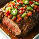Mexican slow cooker beef roast - the image shows a close-up of a cooked steak in a baking dish. the steak appears to be medium-rare and has a dark brown crust on the outside. it is garnished with diced red and green bell peppers, diced tomatoes, and jalapeno peppers. the dish is drizzled with a red sauce, which is visible in the bottom right corner of the image. the background is a white marble countertop.