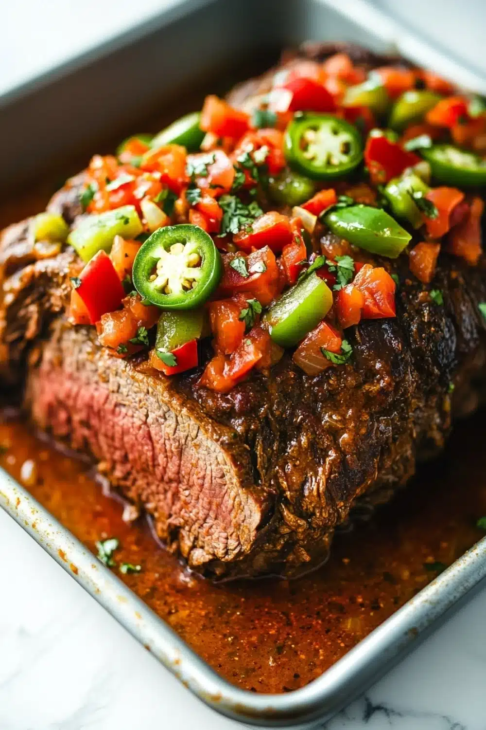 Mexican slow cooker beef roast - the image shows a close-up of a cooked steak in a baking dish. the steak appears to be medium-rare and has a dark brown crust on the outside. it is garnished with diced red and green bell peppers, diced tomatoes, and jalapeno peppers. the dish is drizzled with a red sauce, which is visible in the bottom right corner of the image. the background is a white marble countertop.