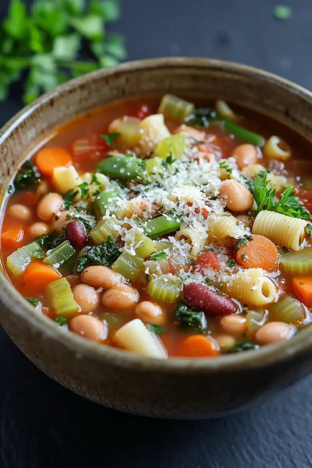 Minestrone soup italian - the image is a close-up of a bowl of soup. the soup appears to be a vegetable soup with white beans, carrots, celery, and other vegetables mixed in. the bowl is made of ceramic and is sitting on a dark grey surface. there is a sprig of fresh parsley on the side of the bowl. the broth is a rich orange color and there are small chunks of red beans scattered throughout the soup. on top of the soup, there is a layer of grated cheese. the overall appearance of the dish is colorful and appetizing.