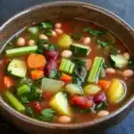 Minestrone soup no pasta - the image is a close-up of a bowl of soup. the soup appears to be a vegetable soup with chunks of white beans, carrots, celery, and other vegetables mixed in. the bowl is made of ceramic and is sitting on a dark grey surface. there are also some sprigs of parsley scattered throughout the soup, adding a pop of green color to the dish. the broth is a reddish-brown color and looks thick and creamy.