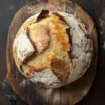 No knead bread dutch oven rise - the image shows a round loaf of bread on a wooden cutting board. the bread appears to be freshly baked, with a golden brown crust and a soft, fluffy interior. the edges of the bread are slightly frayed, indicating that it has been cut in half. the cutting board is made of dark wood and has a rustic, weathered look. there is a small piece of twine hanging from the top right corner of the cutting board, which is likely used to hold the bread in place. the background is a dark, textured surface, possibly a table or countertop.