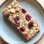 Oatmeal breakfast bars freezer - the image shows a rectangular bar of granola on a light blue plate. the granola appears to be made with oats, nuts, and dried cranberries. the cranberries are bright red and appear to be ripe and juicy. the plate is sitting on a wooden table.