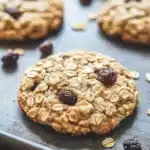 Oatmeal cookie lactation - the image shows a close-up of a freshly baked oatmeal cookie on a baking tray. the cookie is golden brown and has a crumbly texture. it is topped with dried cranberries and scattered nuts. the tray is lined with parchment paper and there are a few more cookies visible in the background. the overall mood of the image is warm and inviting.