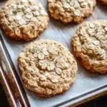 Oatmeal cookies - the image shows a baking tray with freshly baked oatmeal cookies on it. the cookies are round and golden brown in color, with oats scattered throughout. they are arranged in a single layer on the tray, with some overlapping each other. the tray is lined with parchment paper, and the cookies appear to be freshly baked. the background is a wooden table, and there is a dusting of flour on the left side of the tray.