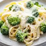 One pot creamy broccoli and parmesan noodles - the image shows a plate of pasta dish with broccoli florets on top. the pasta appears to be fettuccine alfredo, which is a type of italian pasta. the dish is garnished with grated parmesan cheese, which adds a creamy texture and a sprinkle of herbs. the plate is made of light-colored ceramic and is sitting on a white marble countertop. the broccoli is bright green and looks fresh and vibrant.