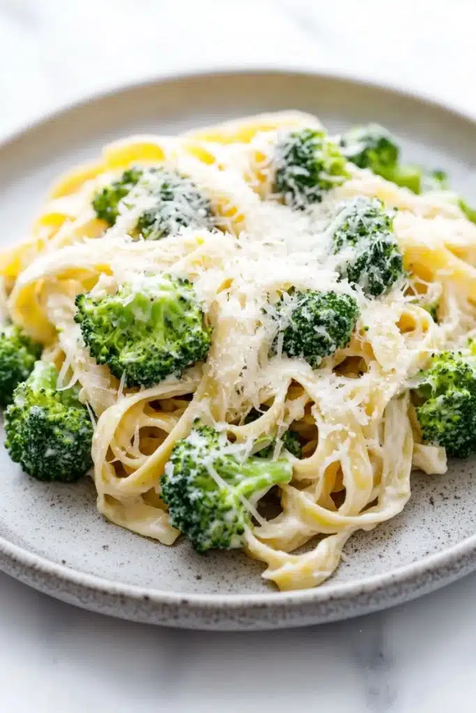 One pot creamy broccoli and parmesan noodles - the image shows a plate of pasta dish with broccoli florets on top. the pasta appears to be fettuccine alfredo, which is a type of italian pasta. the dish is garnished with grated parmesan cheese, which adds a creamy texture and a sprinkle of herbs. the plate is made of light-colored ceramic and is sitting on a white marble countertop. the broccoli is bright green and looks fresh and vibrant.
