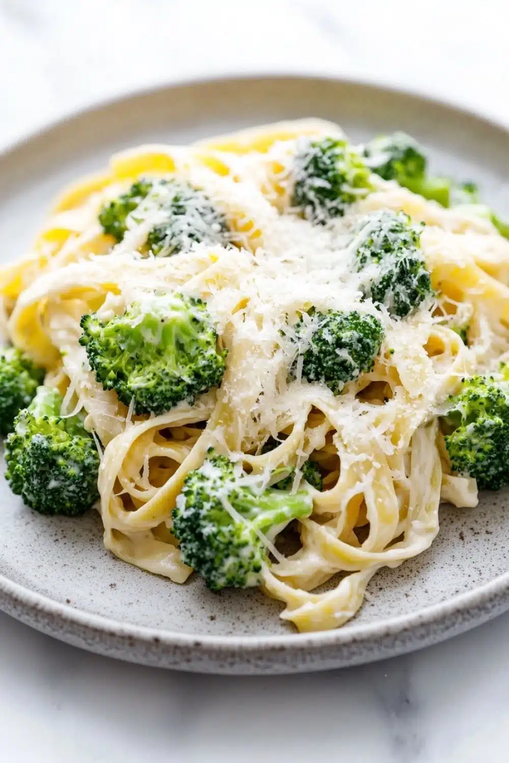 One pot creamy broccoli and parmesan noodles - the image shows a plate of pasta dish with broccoli florets on top. the pasta appears to be fettuccine alfredo, which is a type of italian pasta. the dish is garnished with grated parmesan cheese, which adds a creamy texture and a sprinkle of herbs. the plate is made of light-colored ceramic and is sitting on a white marble countertop. the broccoli is bright green and looks fresh and vibrant.