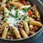 One pot pasta and ground beef - the image is a close-up of a bowl of pasta dish. the pasta appears to be penne, which is a type of penne pasta, and it is cooked in a tomato-based sauce. the dish is garnished with chopped parsley and grated parmesan cheese. the bowl is made of ceramic and is sitting on a dark grey surface. there is a small piece of cheese on the side of the bowl. the background is blurred, but it seems to be a kitchen countertop.