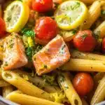 One pot pasta saumon - the image is a close-up of a bowl of pasta dish. the pasta appears to be penne or penne, and it is cooked al dente. on top of the pasta, there are two pieces of salmon, which are cooked to a golden brown color. the salmon is garnished with fresh herbs and lemon wedges. there are also small cherry tomatoes scattered throughout the dish, adding a pop of color and texture. the dish is served in a gray bowl with a white rim. the background is blurred, making the pasta and salmon the focal point of the image.