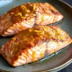 Orange glazed salmon air fryer - the image shows two pieces of salmon on a green plate. the salmon is cooked to a golden brown color and appears to be seasoned with herbs and spices. the plate is sitting on a wooden table with a dark background. the food is arranged neatly on the plate, with the salmon on the left side and the one on the right side. the sauce on the salmon is a golden yellow color and is drizzled over the top of the salmon. the overall appearance of the dish is elegant and appetizing.